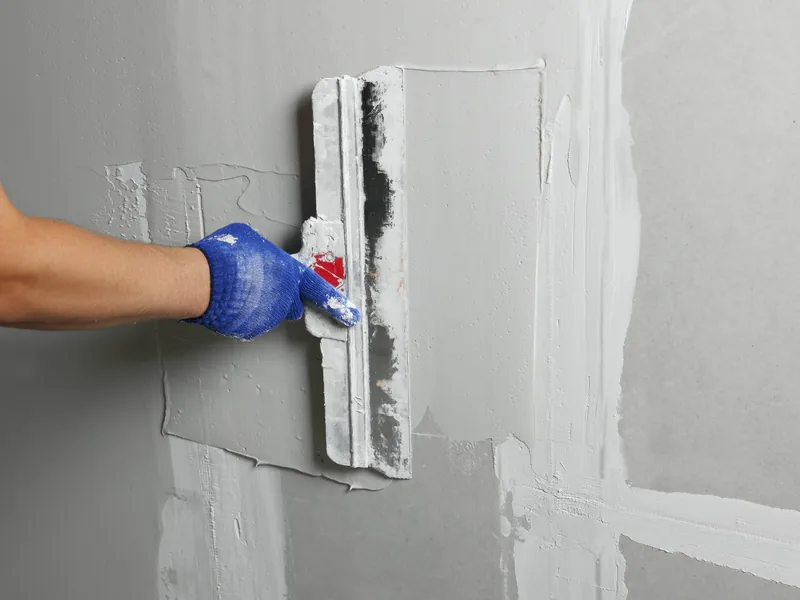 A gloved hand drawing a wide knife of compound across a patched wall