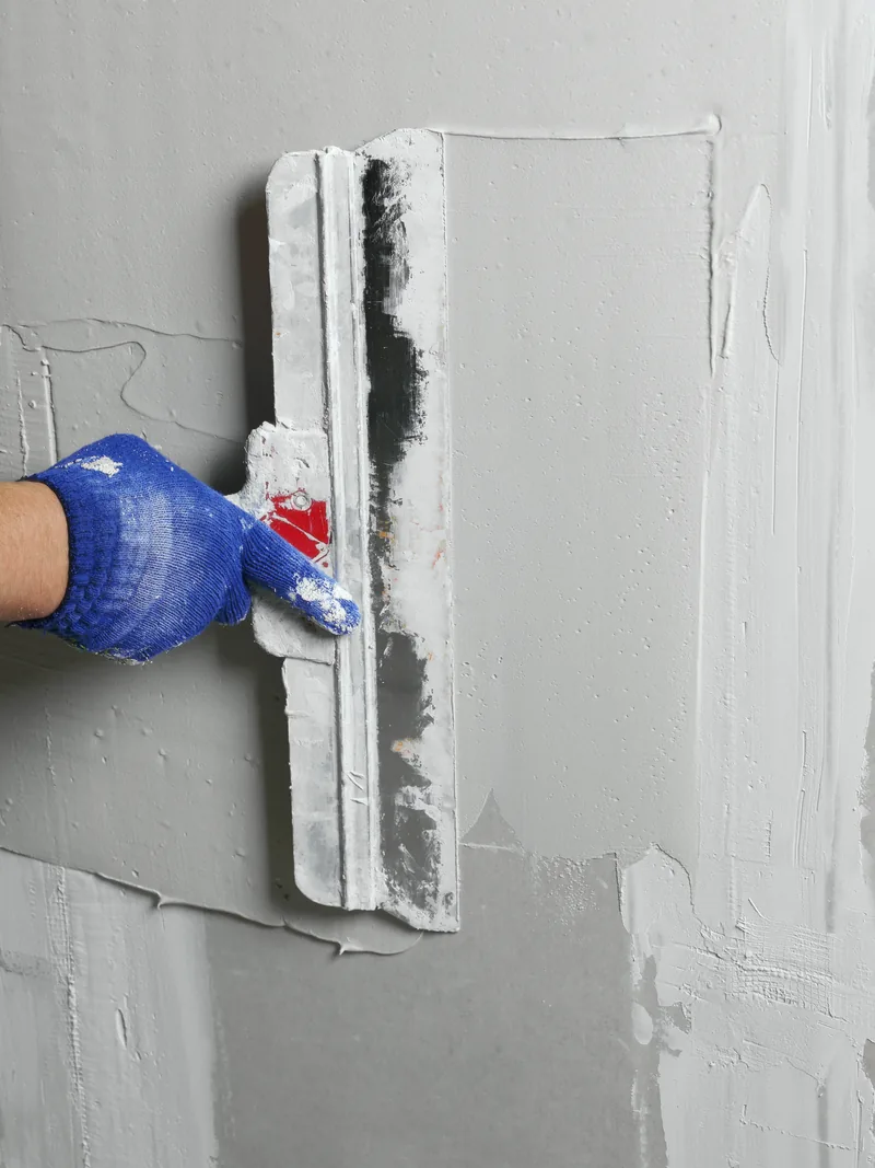 A gloved hand drawing a wide knife of compound across a patched wall