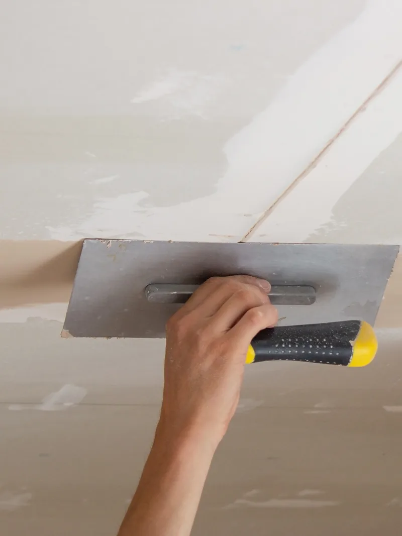 A hand flattening compound across an overhead ceiling seam