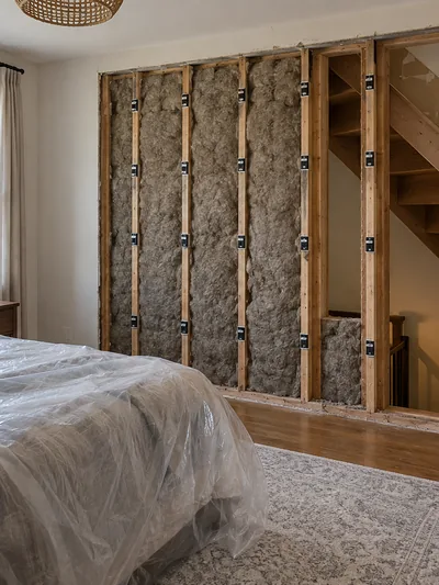 A finished bedroom wall opened to bare studs and insulation beside a stairwell