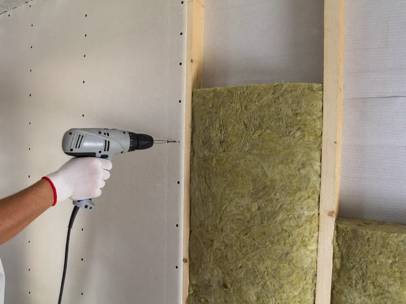 A screw gun driving a fastener through board beside an open insulated bay
