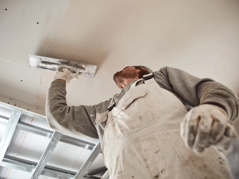 A worker reaching overhead with a knife at a suspended ceiling frame