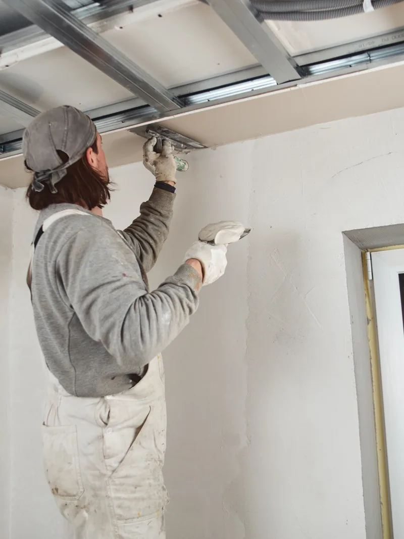 A worker finishing a wall beside a work light angled across the surface