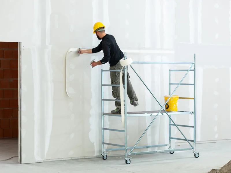 A worker on a rolling tower taping the upper part of a tall wall