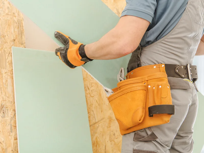 Gloved hands lowering a green moisture-resistant panel onto a sheathed wall