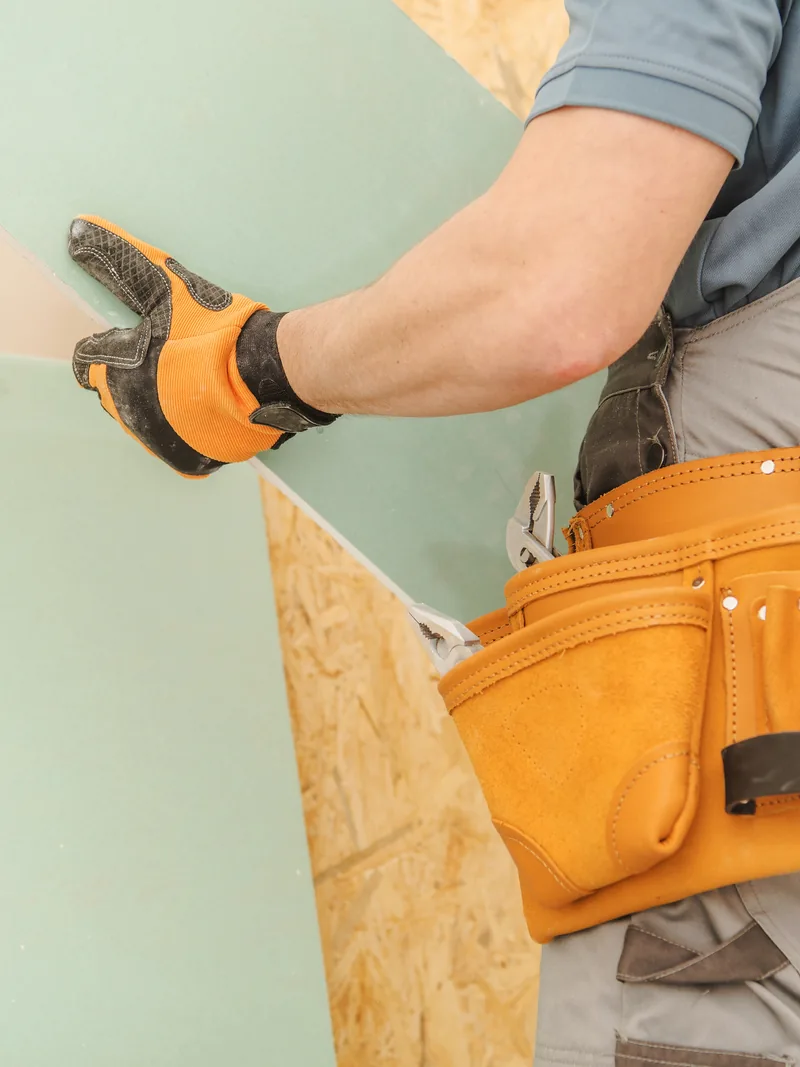 Gloved hands lowering a green moisture-resistant panel onto a sheathed wall