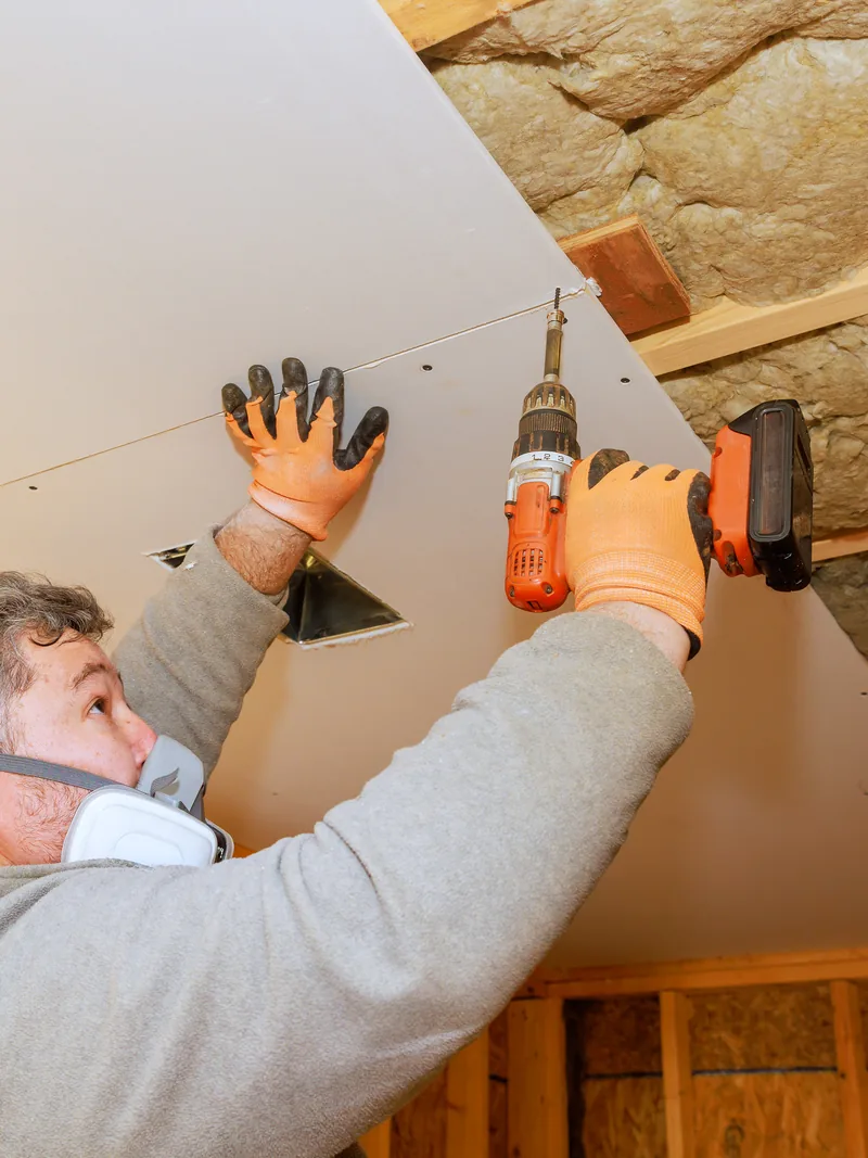 A worker driving screws into a ceiling panel between insulated joists