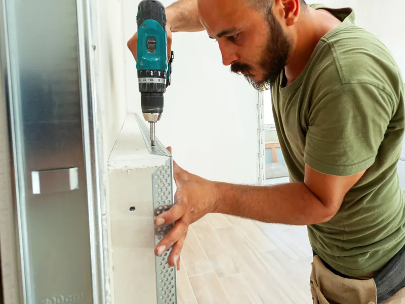 A worker fixing a metal corner bead to an outside corner with a drill
