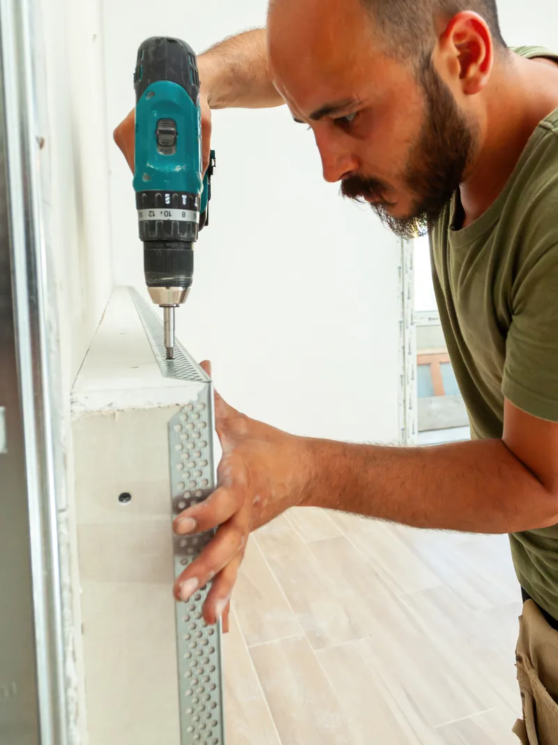 A worker fixing a metal corner bead to an outside corner with a drill