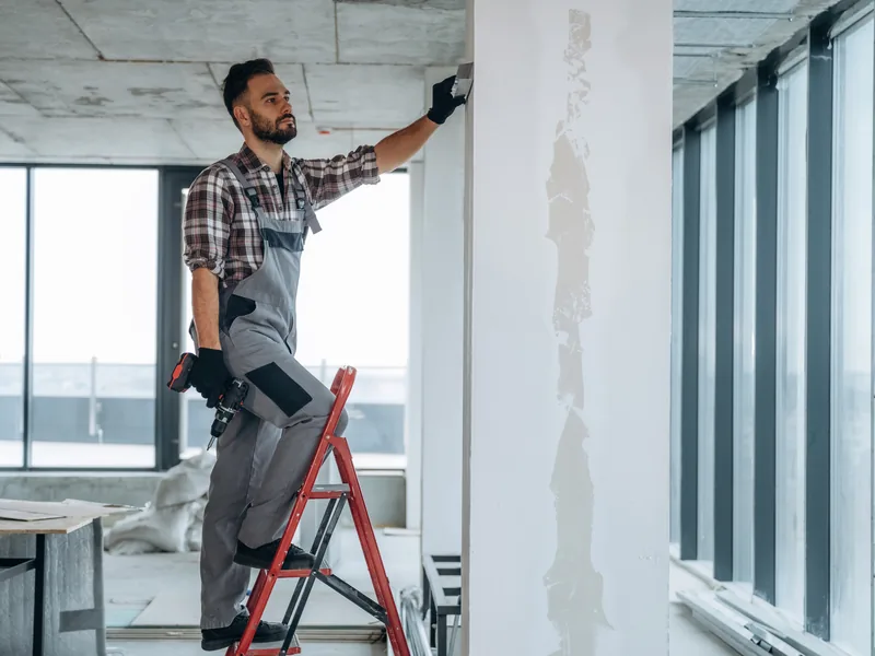 A worker on a stepladder finishing a wall beside a glazed office partition