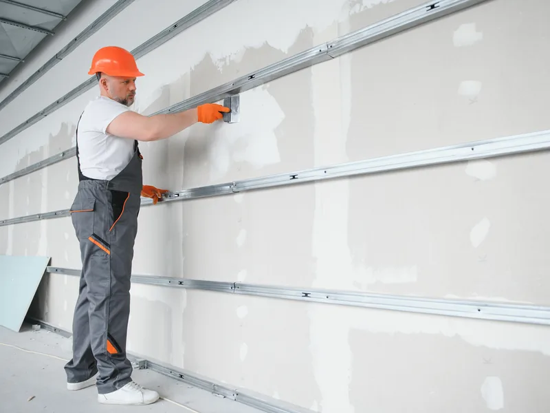 A worker running a knife along a wall lined with horizontal steel furring