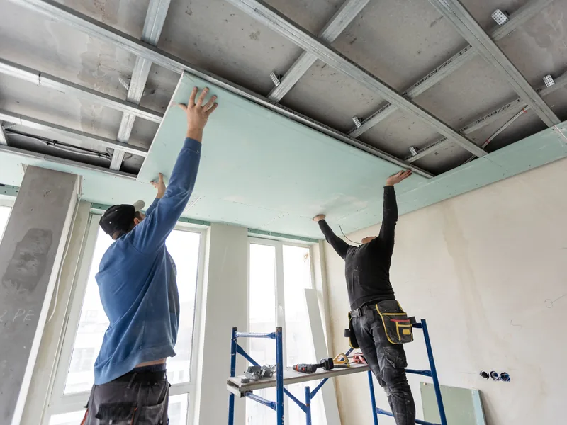 Two workers fitting panels into an exposed ceiling grid from step platforms
