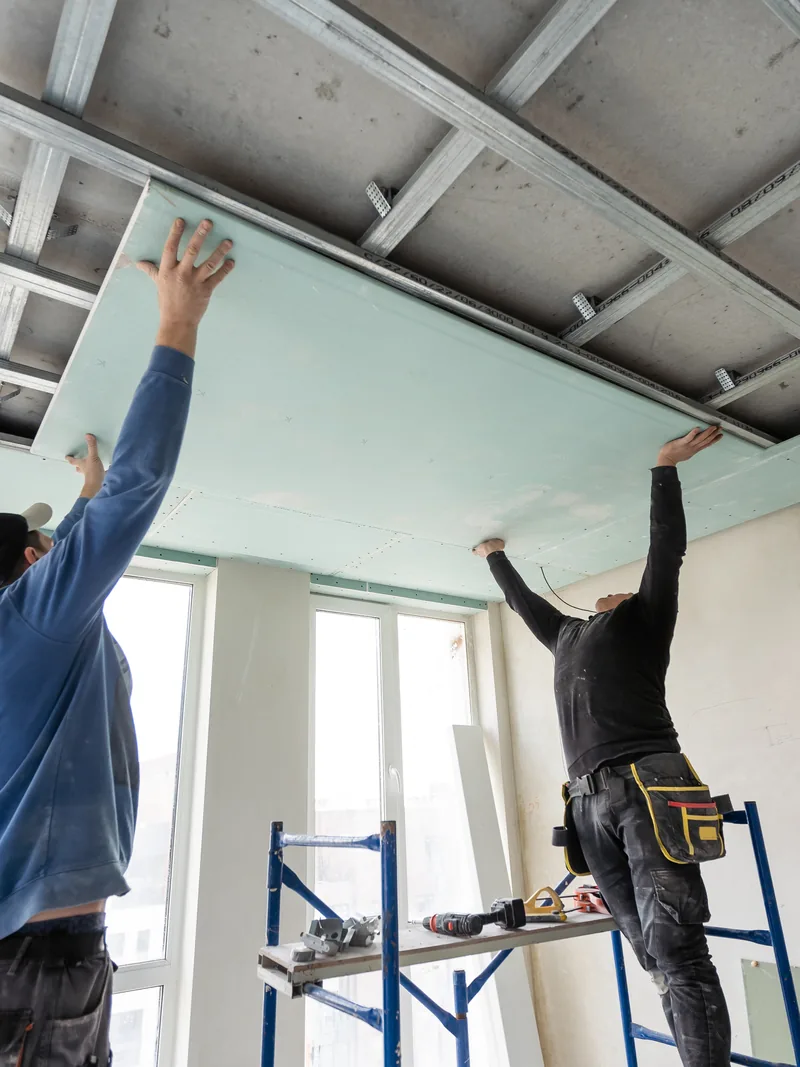 Two workers fitting panels into an exposed ceiling grid from step platforms