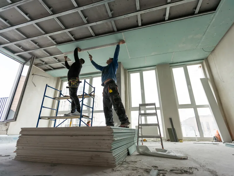 Two workers on a platform lifting a green panel to a ceiling frame