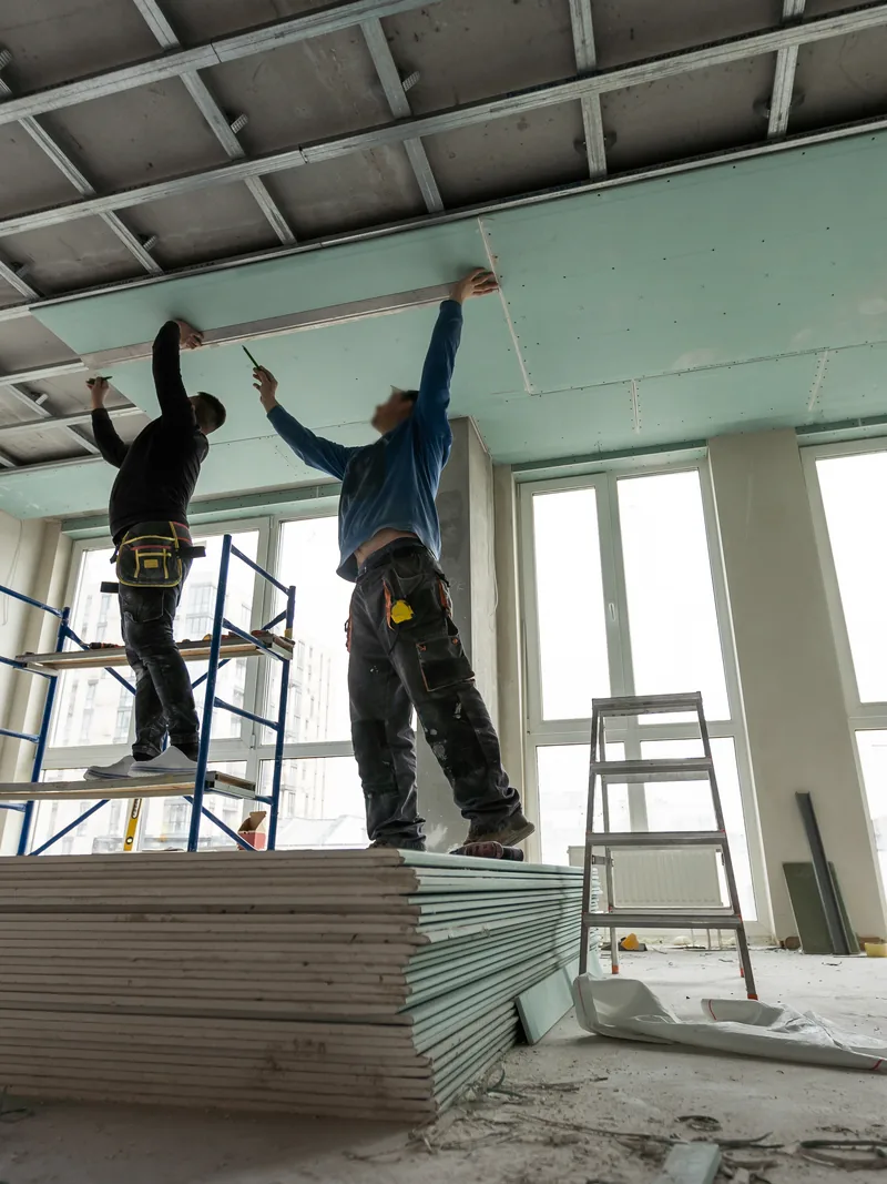 Two workers on a platform lifting a green panel to a ceiling frame