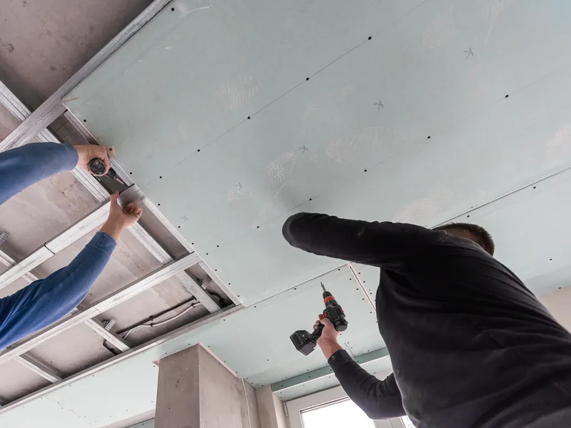 A worker drilling into a boxed ceiling drop with ductwork running above it