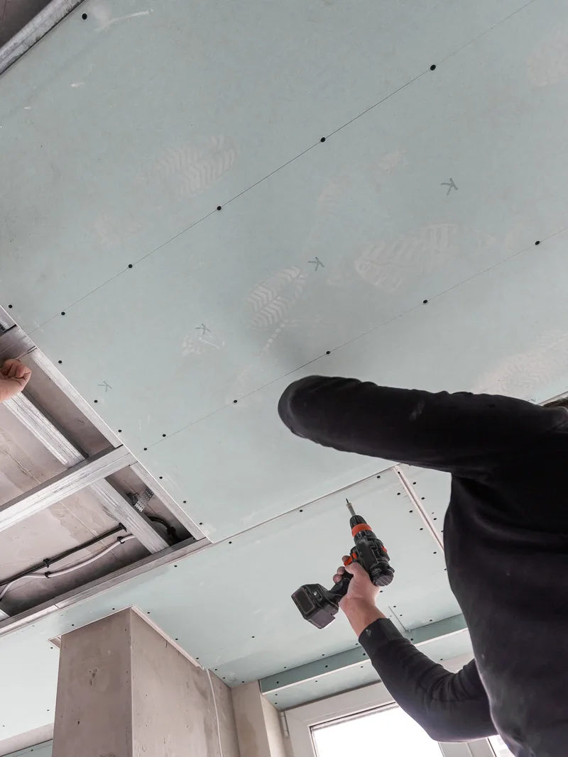 A worker drilling into a boxed ceiling drop with ductwork running above it