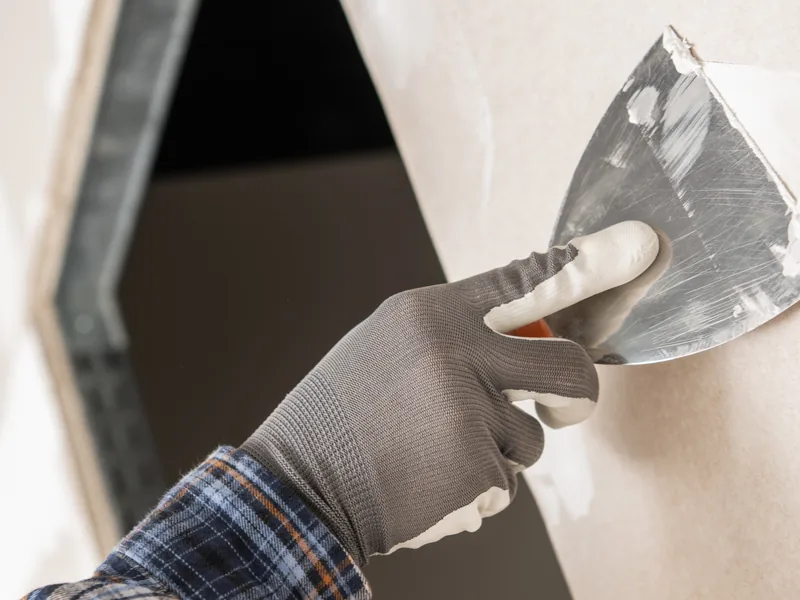 A gloved hand carrying a trowel of compound to a dark wall opening