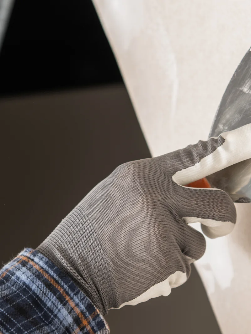 A gloved hand carrying a trowel of compound to a dark wall opening